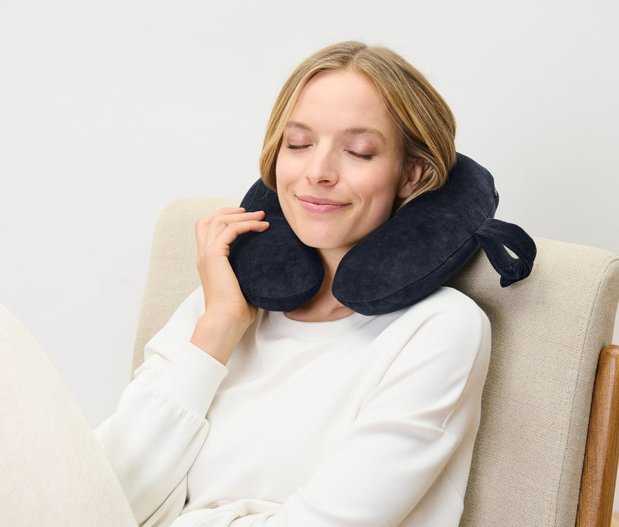 Egy nő csukott szemmel ül egy széken, és egy sötétkék nyakpárnát visel.