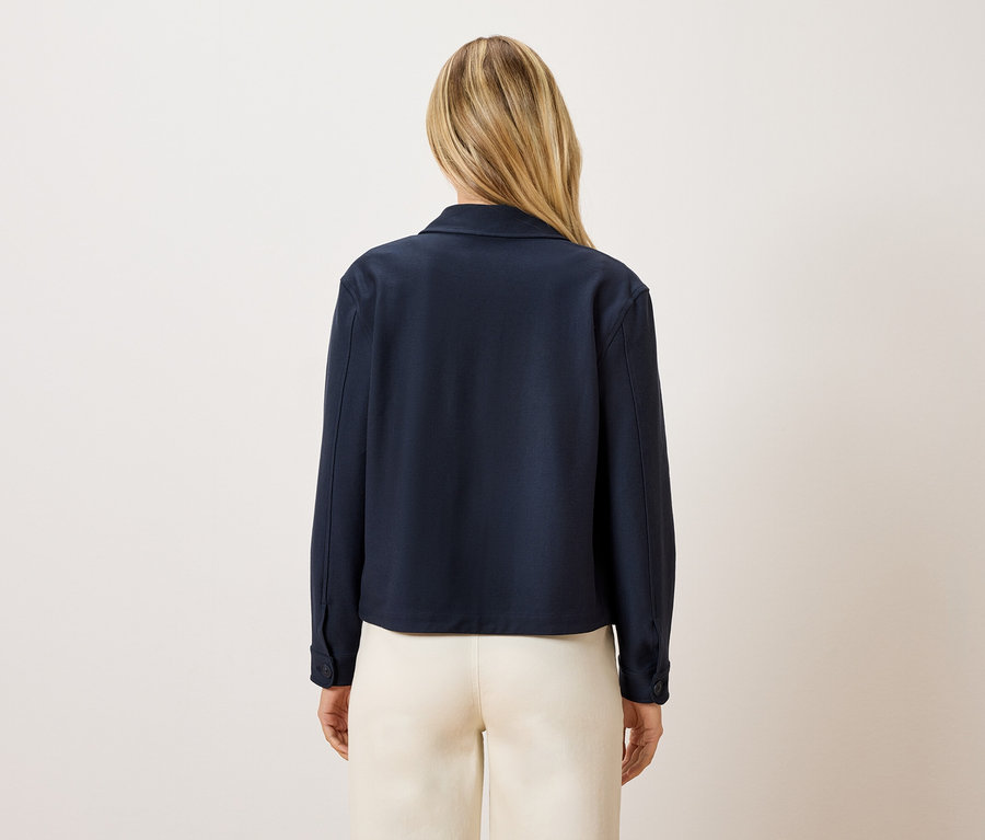 Hátulnézet egy szőke hajú nőről, aki sötétkék kabátot és krémszínű nadrágot visel.