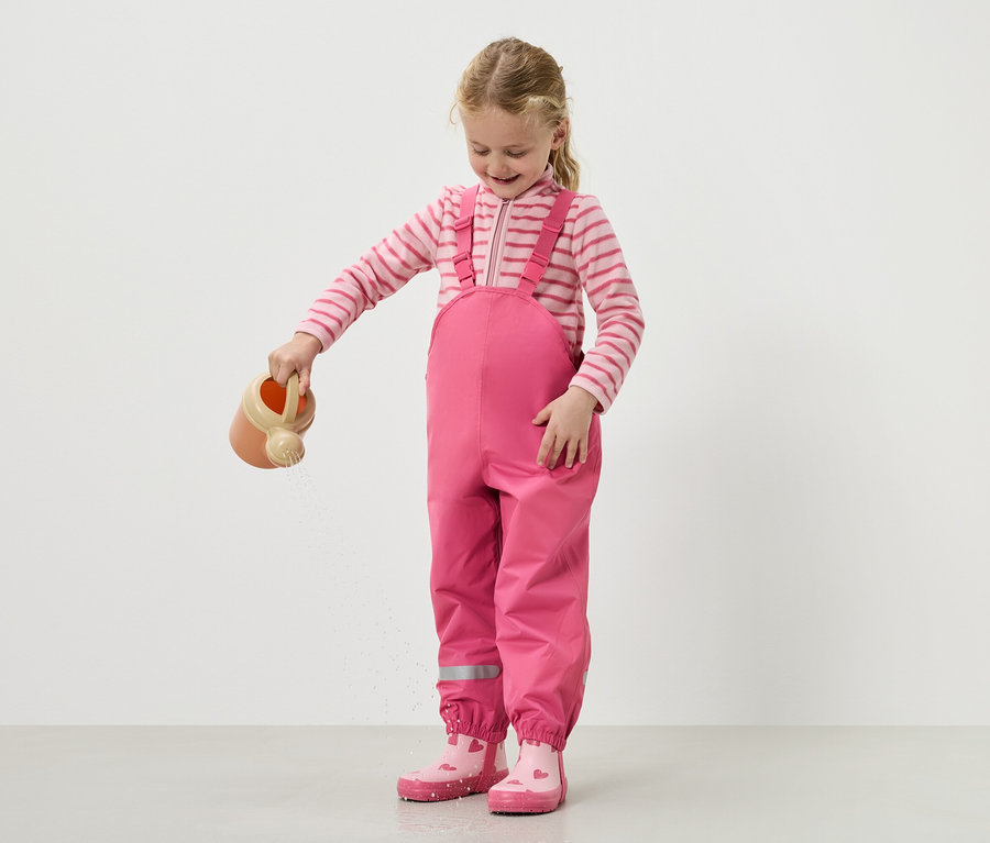 Egy lány rózsaszín kertésznadrágban és csizmában locsolja a vizet egy locsolókannából.