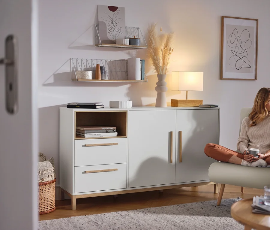 Egy nő ül a nappaliban egy fehér, fiókos és ajtós »Eklund« komód mellett.