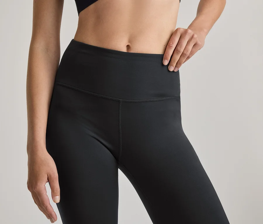 Közeli felvétel egy személy törzséről, aki fekete magas derekú leggingset és fekete sportmelltartót visel. Jobb keze a csípőjén van.