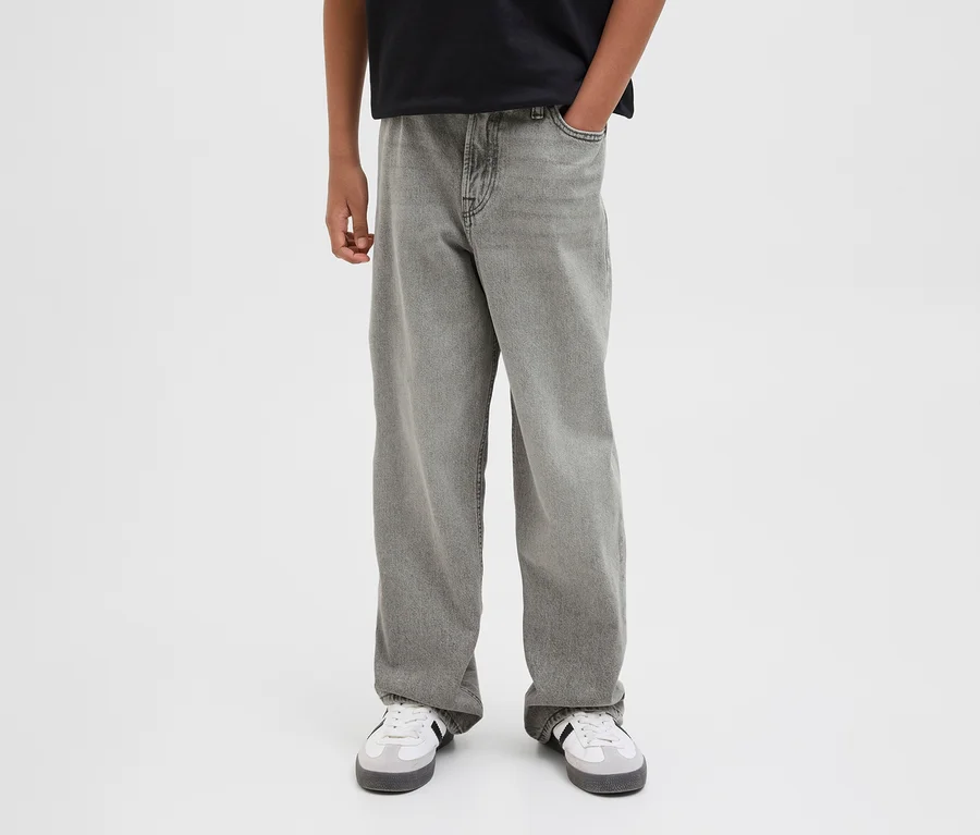 Egy személy szürke Jack&Jones JJOrginial Alex farmernadrágot és fehér sportcipőt visel. Egyik keze a zsebében van.