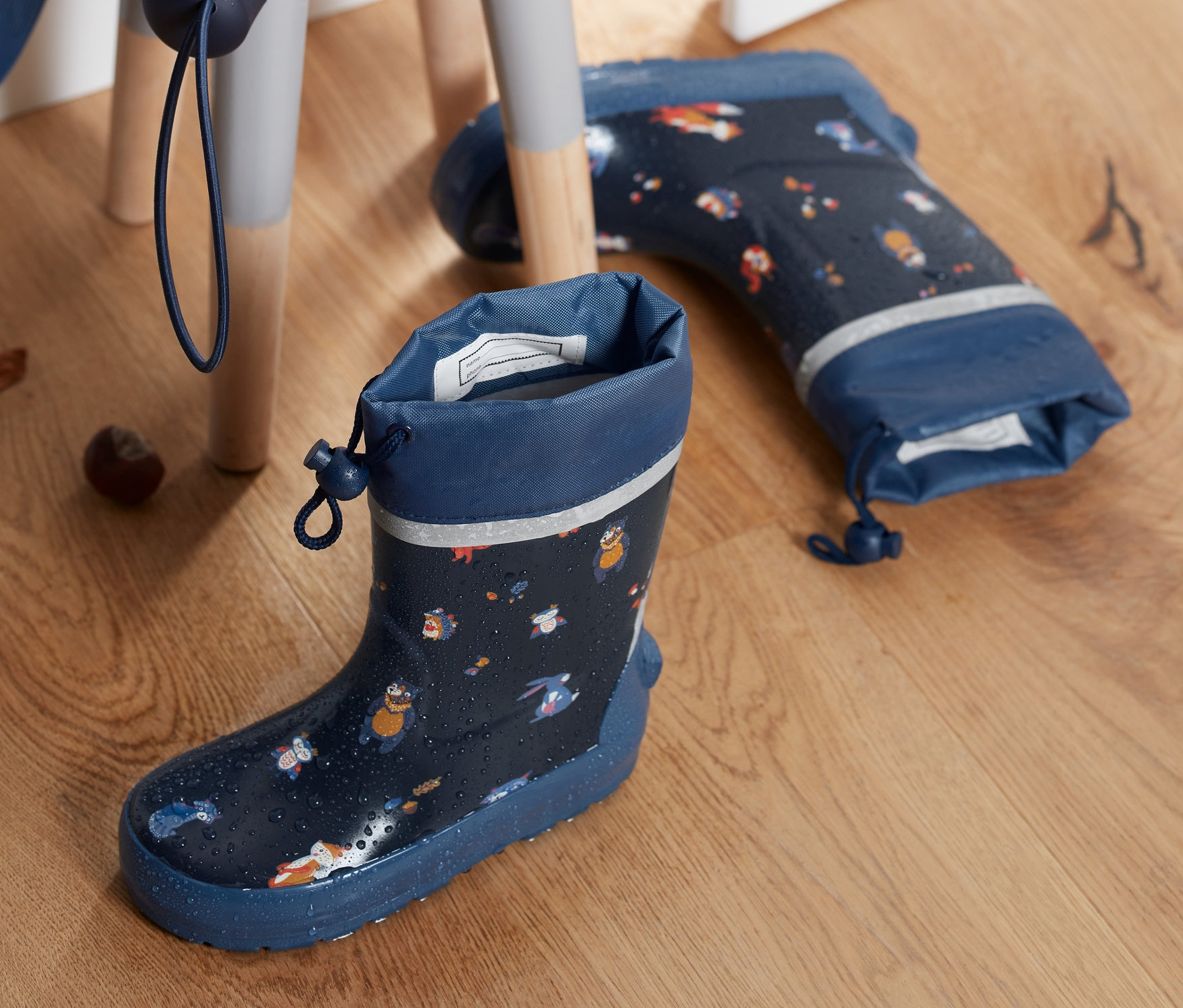 Sötétkék, teljesen nyomott mintás gumicsizmák egymás mellett a fapadlón, egy összecsukott esernyő és gesztenyék egy széken.