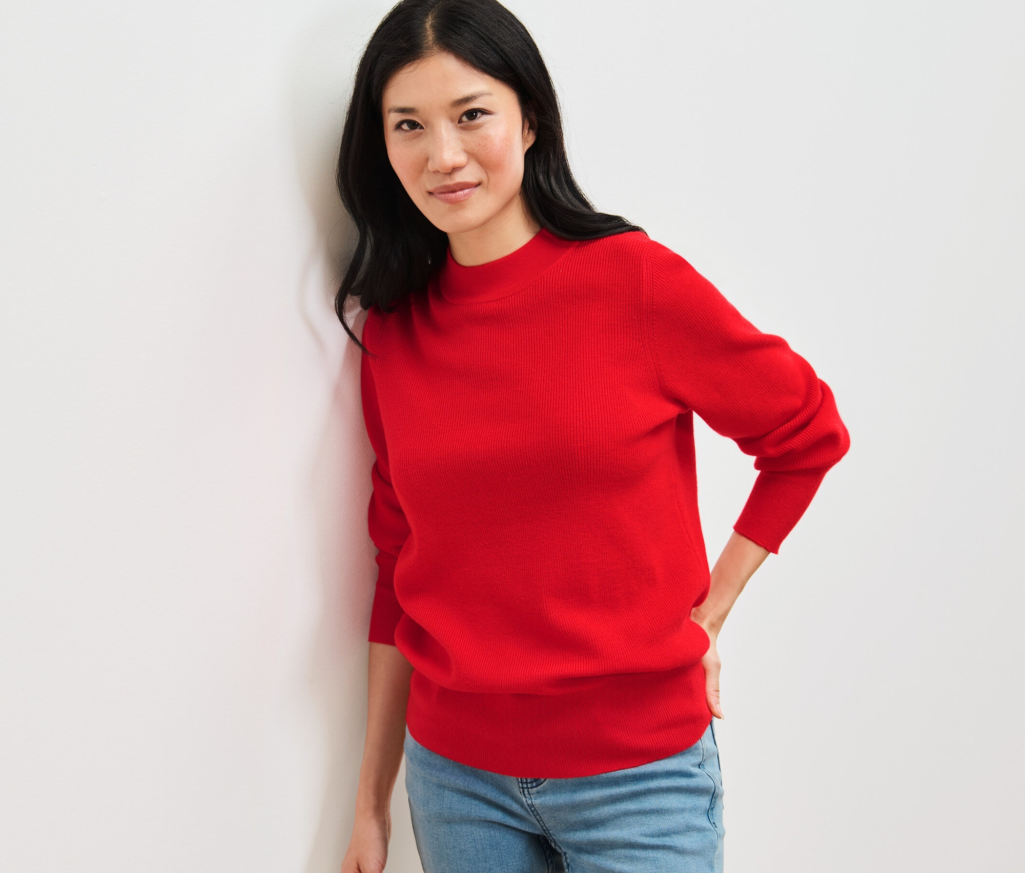 Egy nő a falnak támaszkodik, piros, finomkötésű pulóvert és farmert visel.