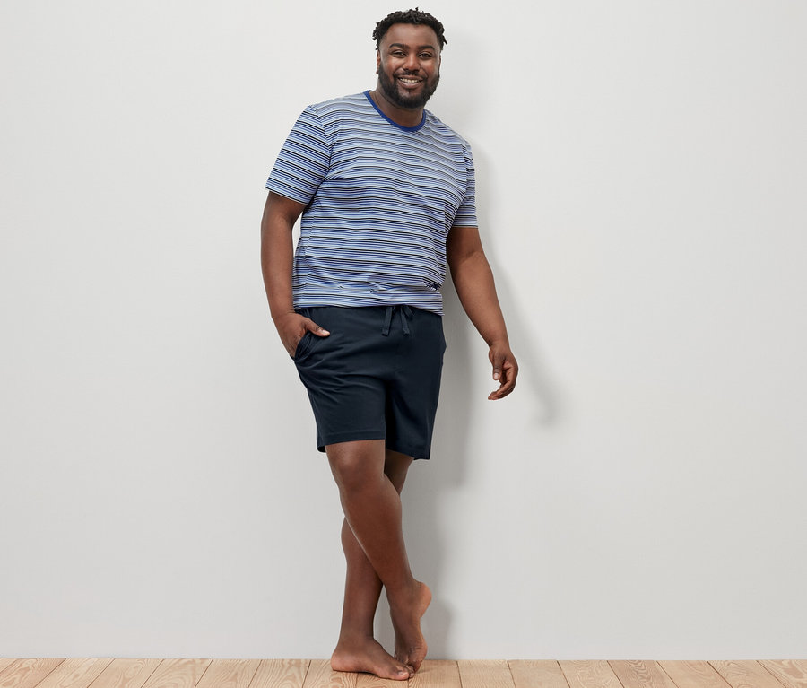 Férfi pózol a fa padlón, kék-sötétkék csíkos rövidnadrágos pizsamában.