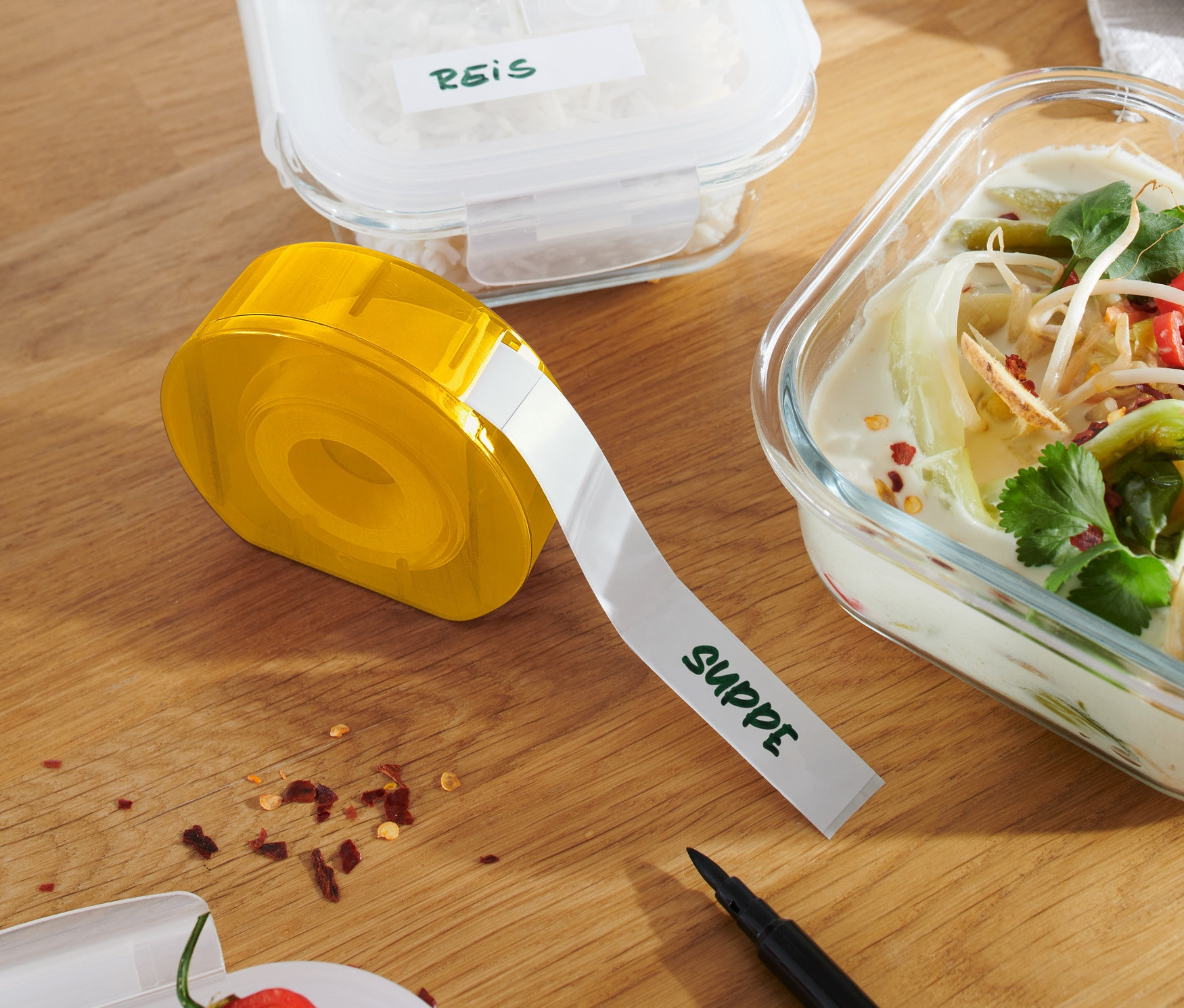 Sárga címkeadagoló egy fa asztalon, rizzsel és levessel teli edények mellett.