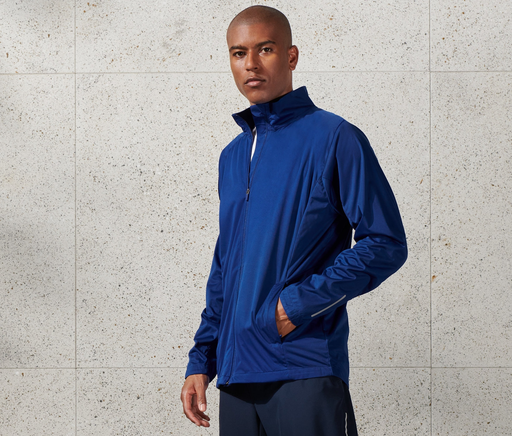 Férfi kék szélálló funkcionális dzsekit, rövidnadrágot és fehér tornacipőt visel.