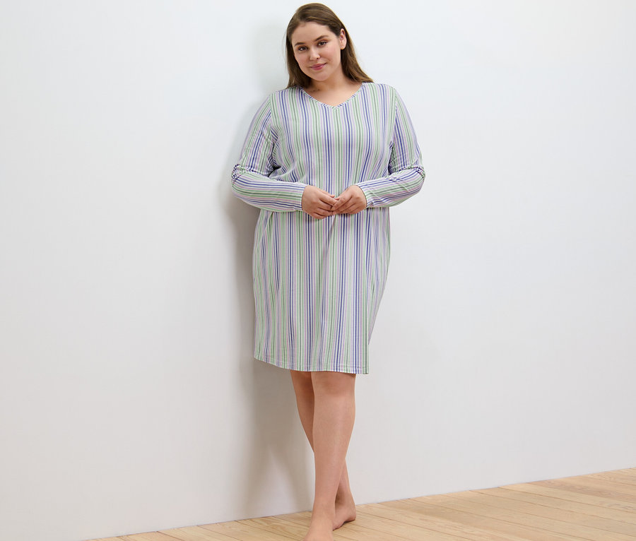 Nő csíkos hálóingben áll a falnál.