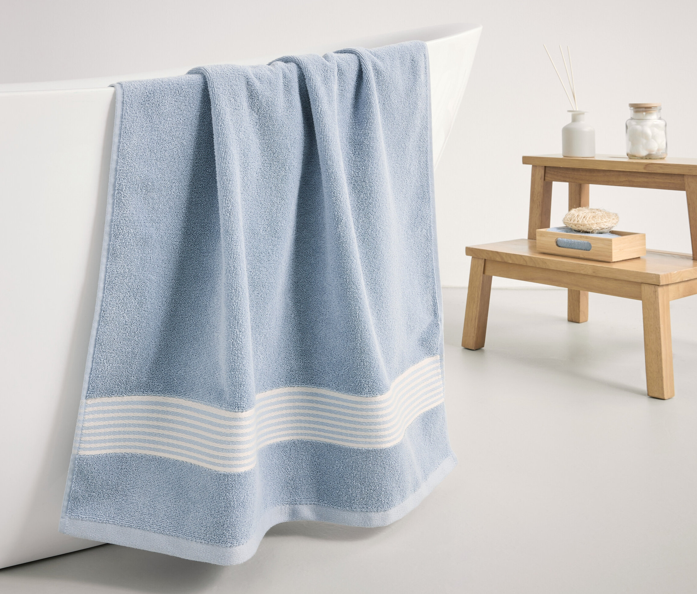 Egy világoskék prémium fürdőlepedő lóg egy fehér fürdőkád szélén.