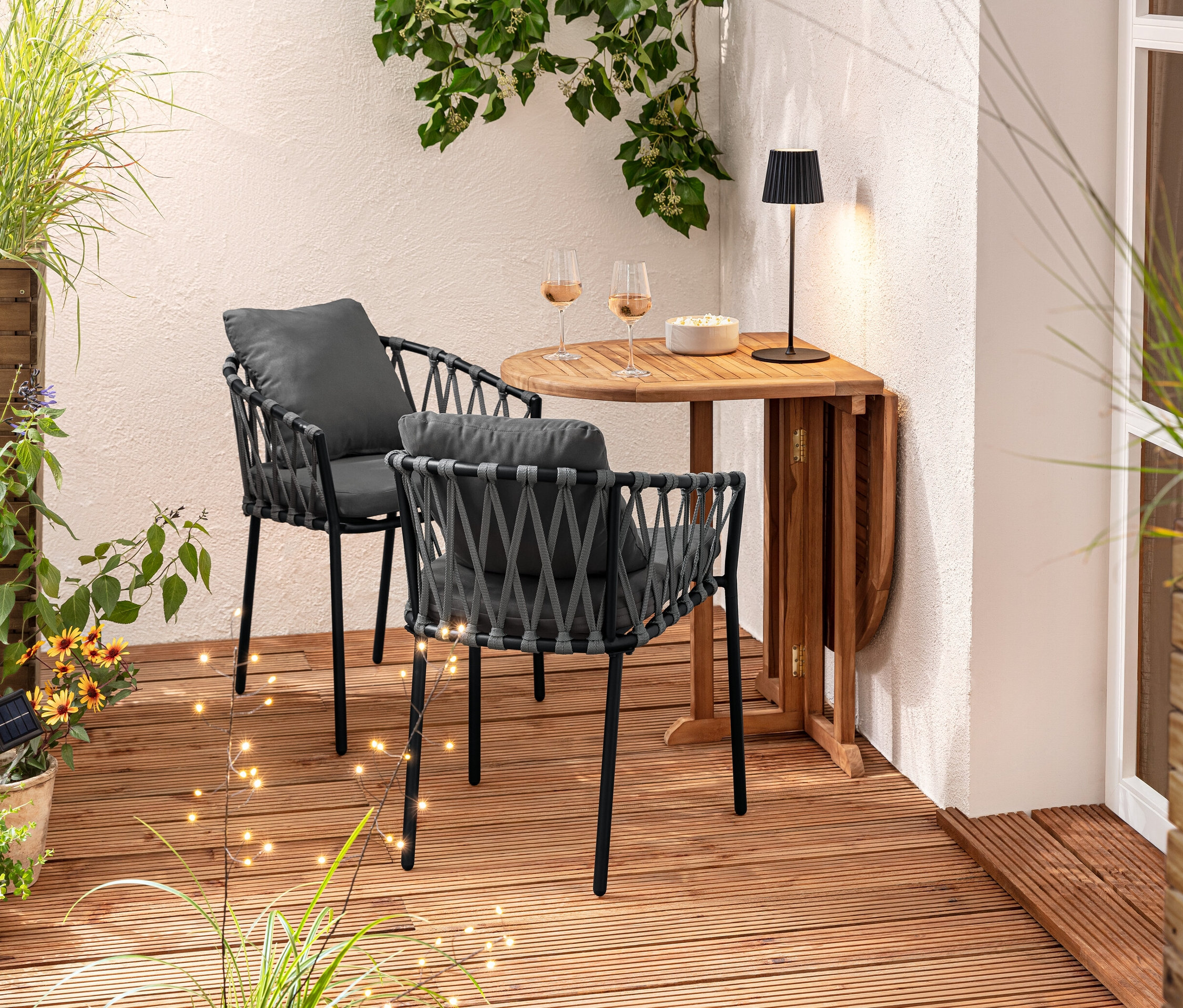 Két textilhálós étkezői karosszék áll egy fa asztalnál. Az asztalon poharak és egy tál látható. A háttérben egy »Tinus« térelválasztó virágláda és 3 szolárnyárs található.