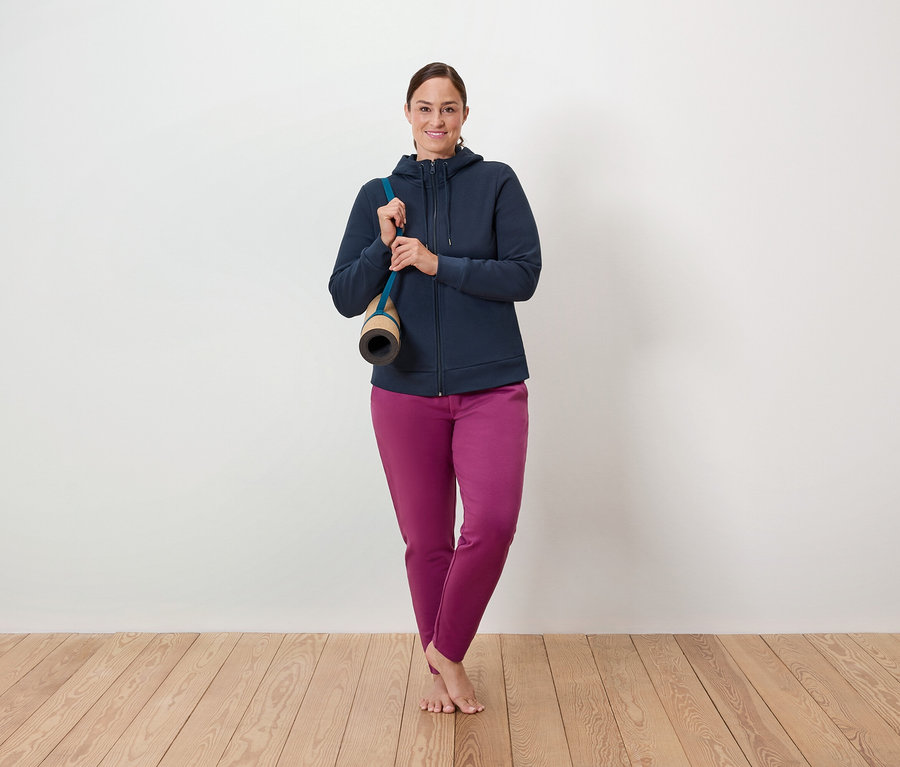 Egy nő áll egy fapadlón, a vállán egy jógaszőnyeg, kék cipzáras pulóvert és lila nadrágot visel.