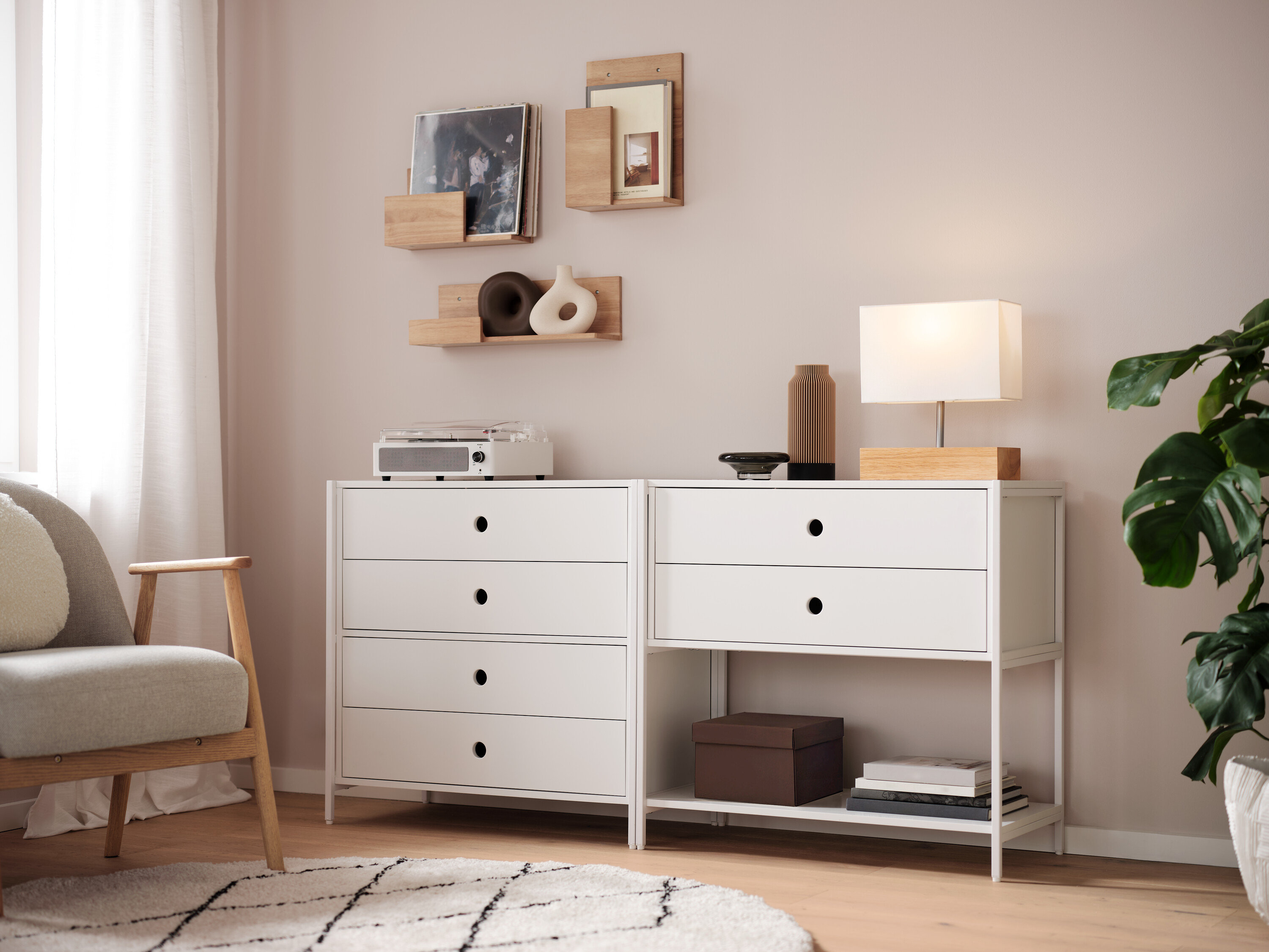 Két fehér komód áll egymás mellett, rajtuk egy lámpa, egy váza és egy rádió. A falon polcok vannak.