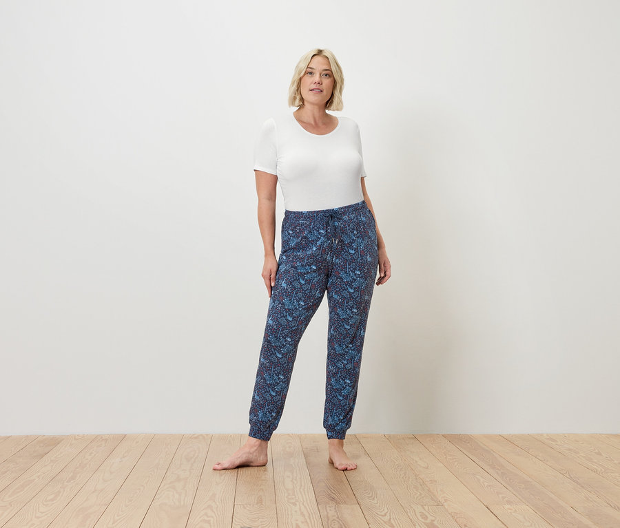 Egy nő mezítláb áll a fapadlón, fehér pólót és kék jogging nadrágot visel.