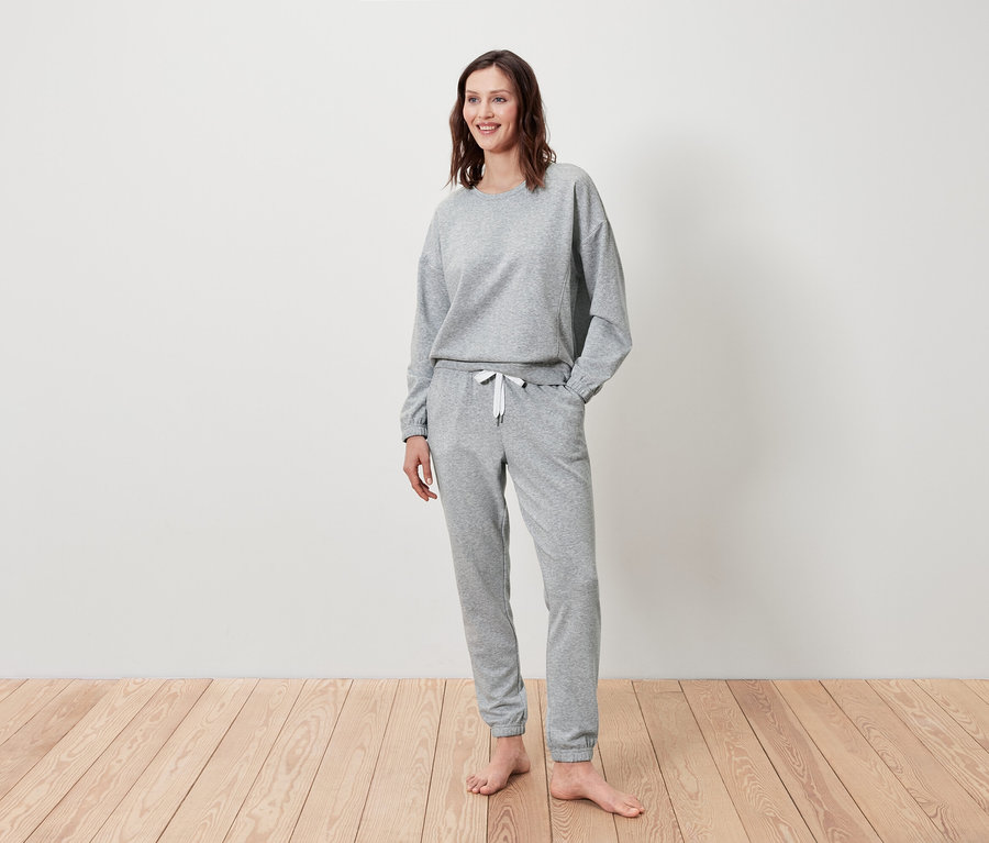 Egy nő áll és mosolyog, szürke pulóvert és melegítőnadrágot visel.