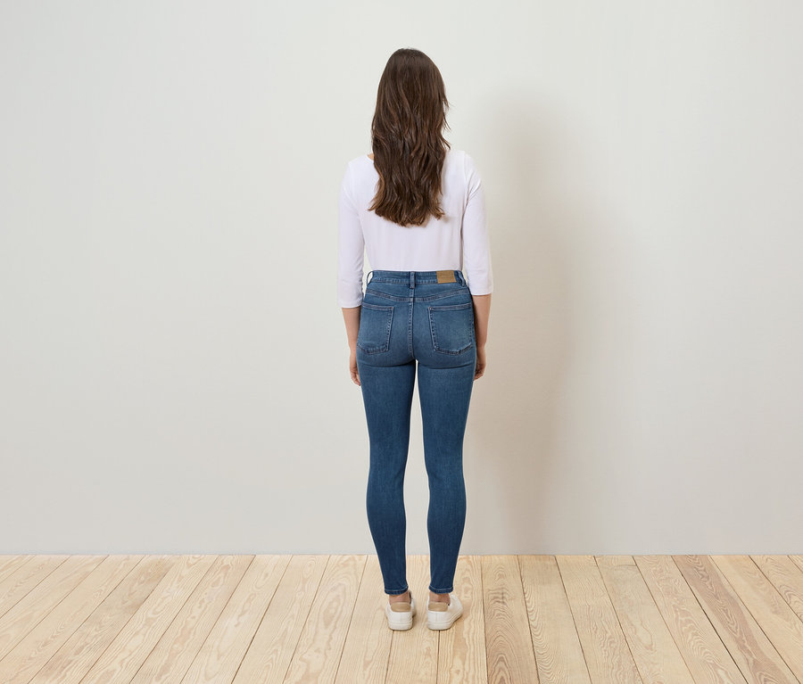 Egy nő áll háttal a kamerának, kék farmernadrágot és fehér blúzt visel.