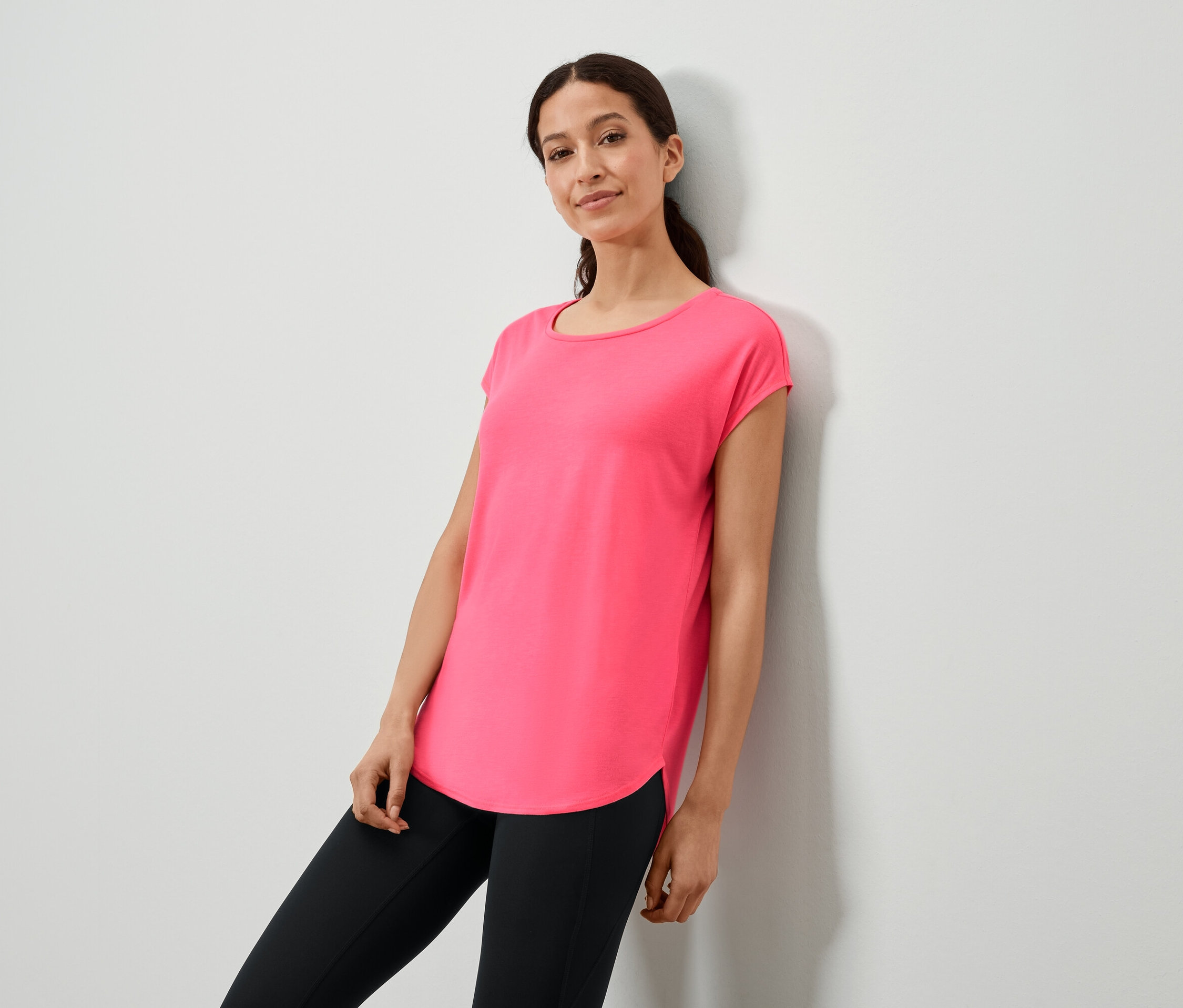 Nő támaszkodik a falnak rózsaszín pólóban és fekete leggingsben.