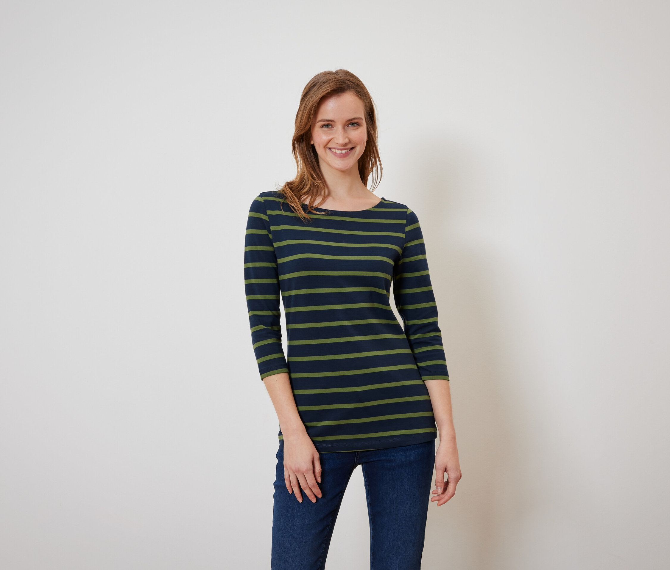 Barna hajú nő pózol kék-zöld csíkos pólóban és kék farmerben.