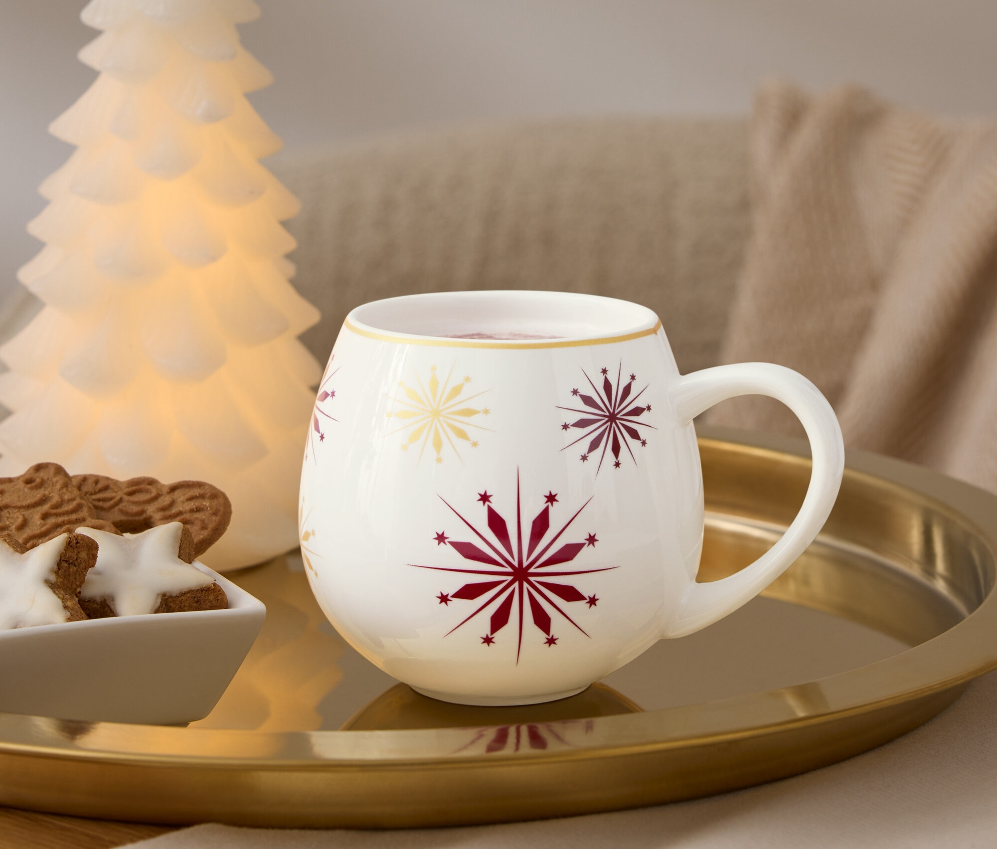 Egy arany tálcán egy piros és arany hópelyhekkel díszített fehér bögre, egy mézeskalácsot tartalmazó tál és egy világító fa áll.