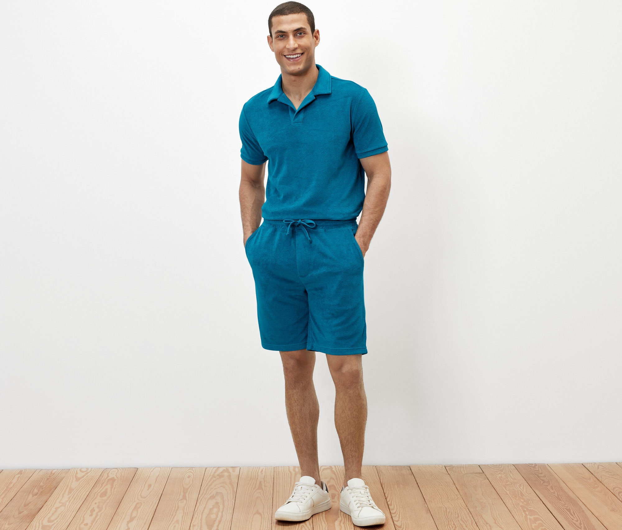 Egy férfi áll, kezével a petrolkék frottír rövidnadrágjának zsebében.