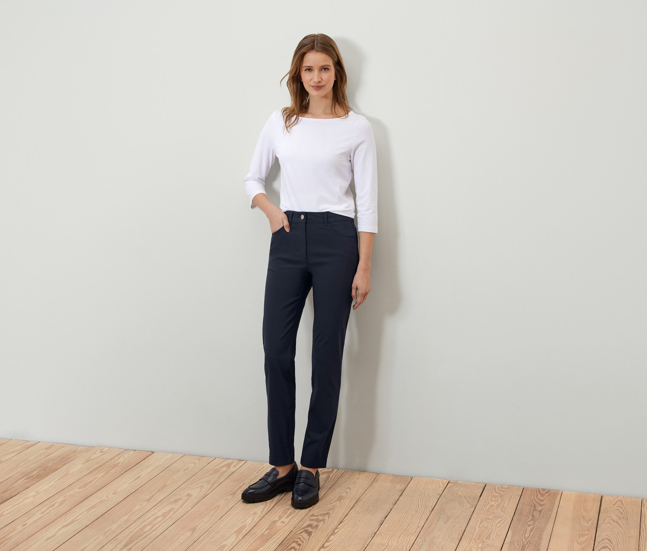 Egy nő áll és pózol egy fehér háromnegyedes ujjú pólóban és sötétkék nadrágban.