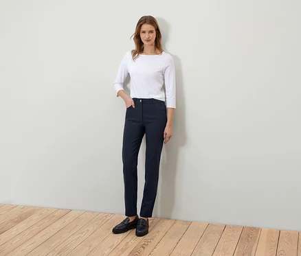 Egy nő áll és pózol egy fehér háromnegyedes ujjú pólóban és sötétkék nadrágban.
