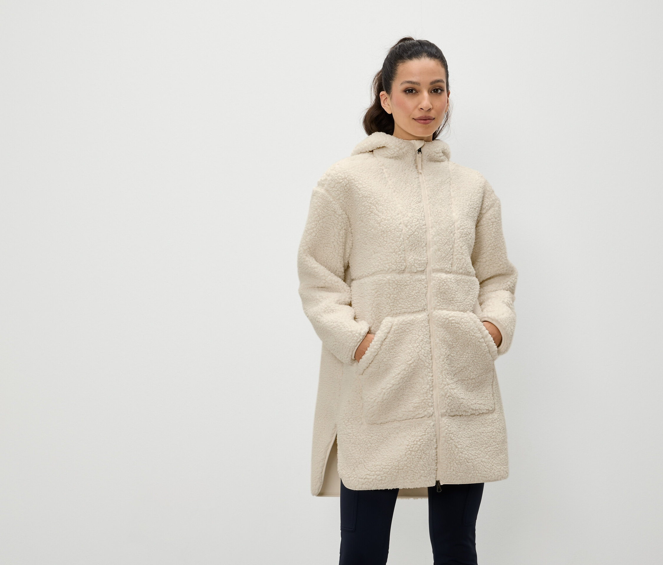 Egy nő áll, kezével egy krémszínű, cipzáras, kapucnis bélelt kabát zsebében.