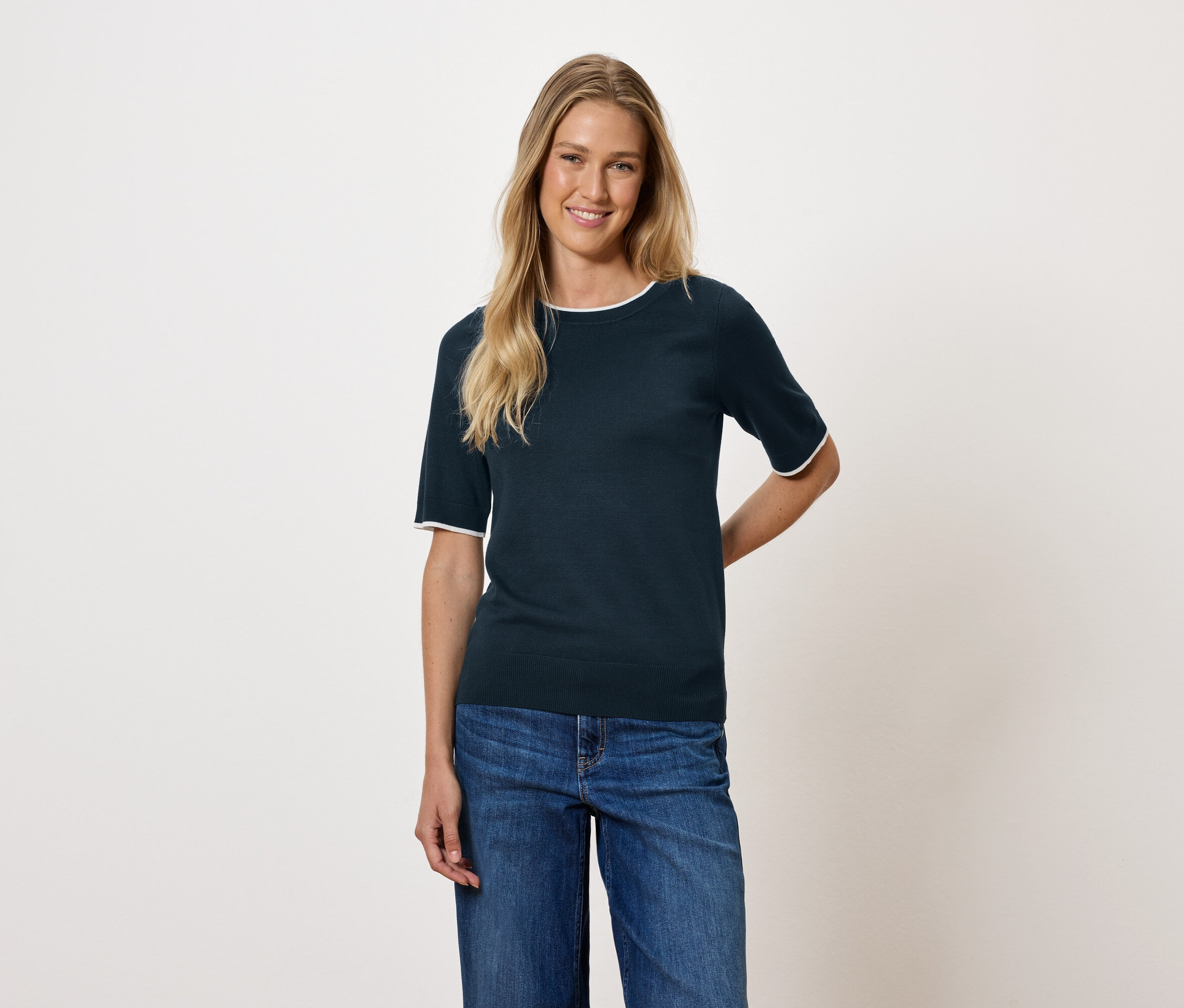 Szőke hajú nő sötétkék rövid ujjú felsőt és farmert visel.