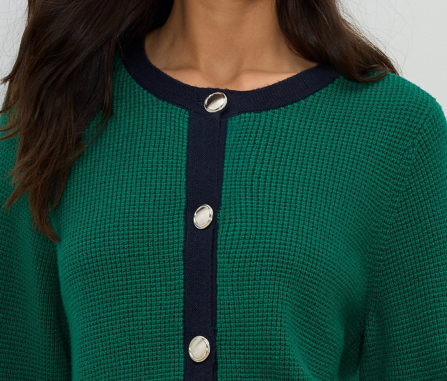Közeli kép egy zöld kötött kardigánt viselő nőről, sötétkék részletekkel és ezüst gombokkal.