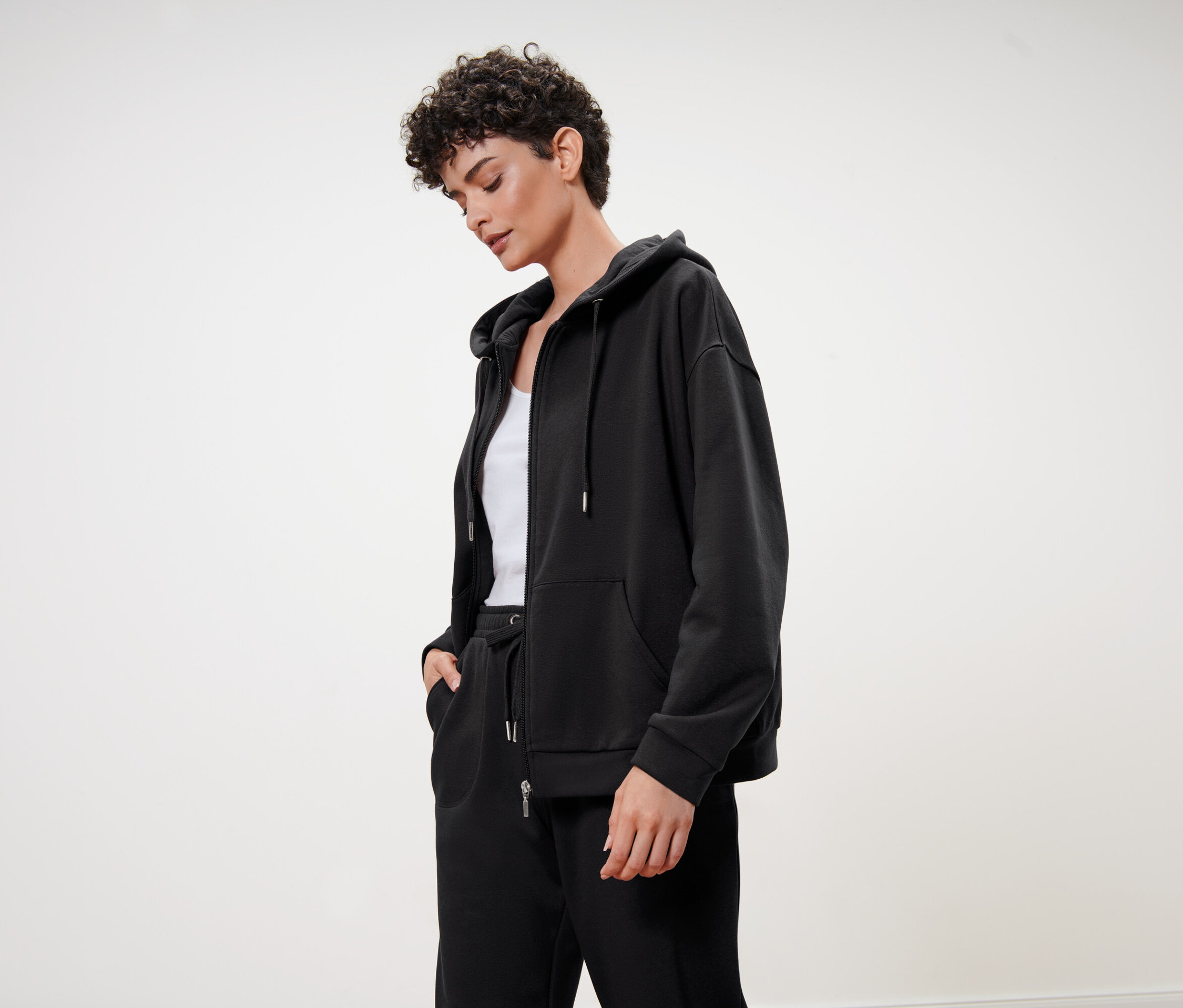 Rövid, göndör hajú nő fekete kapucnis pulóvert, fekete melegítőnadrágot és fehér felsőt visel.