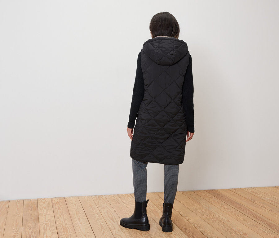 A nő háttal áll a kamerának, steppelt fekete mellényt visel kapucnival, fekete csizmát és szürke leggingset.