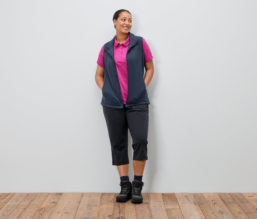 Egy nő áll a kezével a zsebében egy fal előtt. Tengerészkék mellényt visel egy rózsaszín póló felett, fekete nadrágot és csizmát.