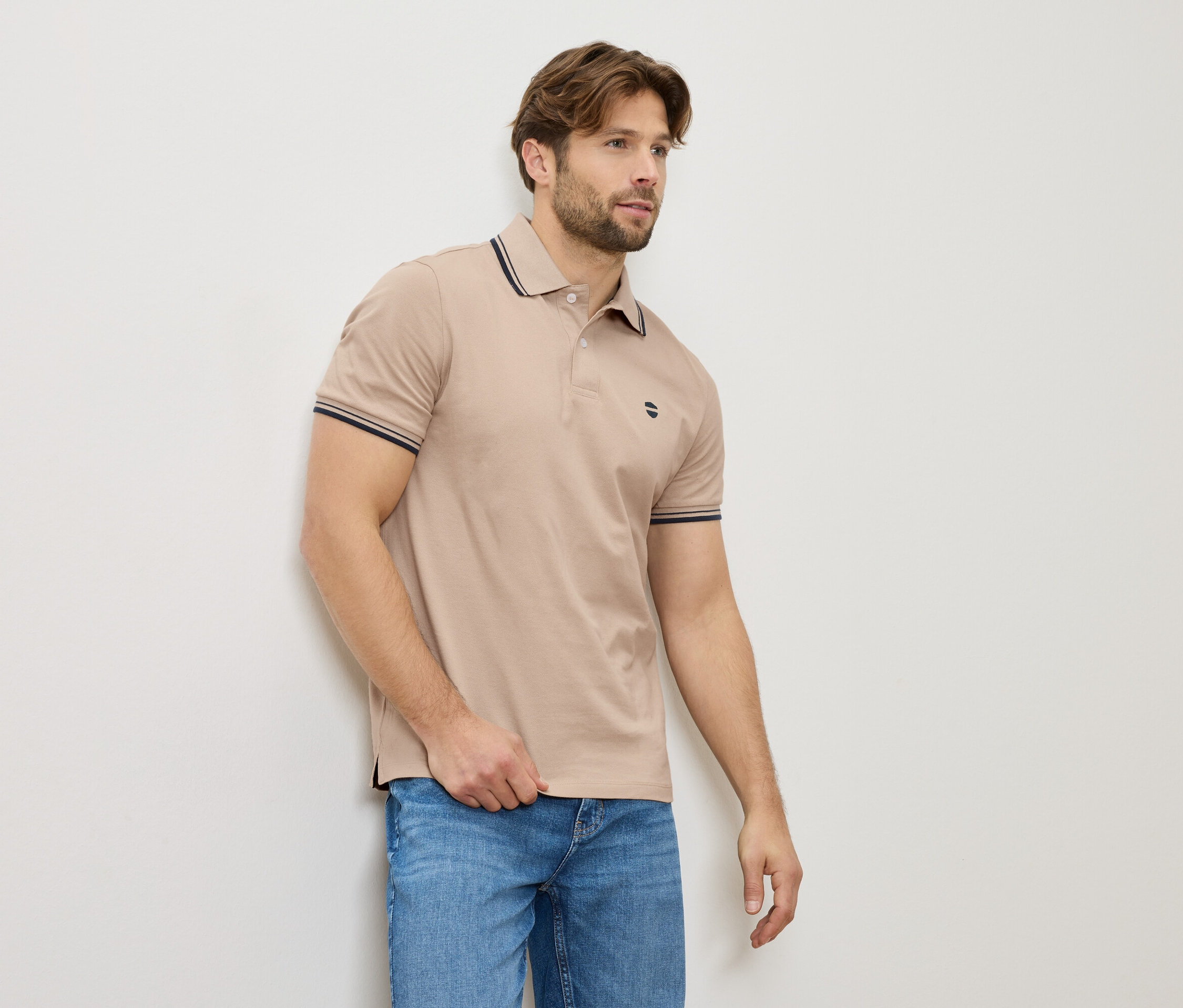 Egy férfi bézs színű pólóban és farmerben áll a falnál.