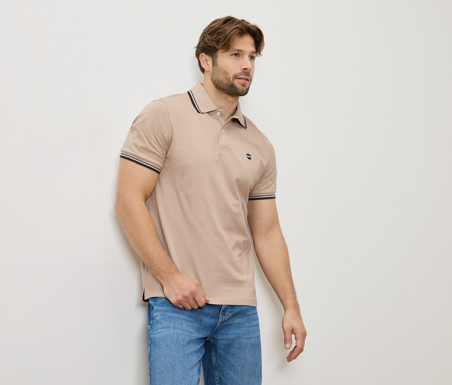 Egy férfi bézs színű pólóban és farmerben áll a falnál.