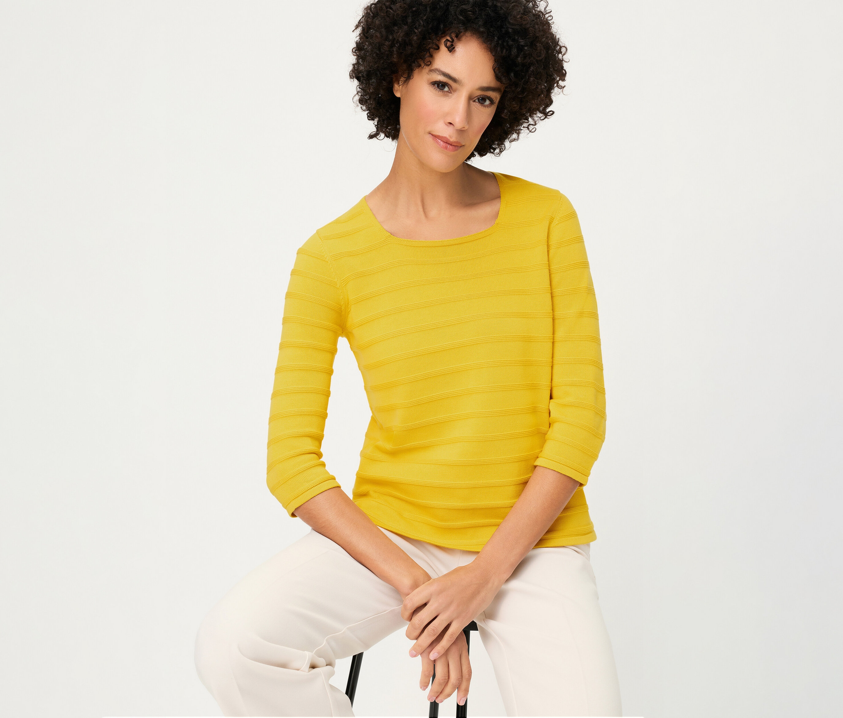 Egy nő ül, és egy sárga, strukturált, 3/4-es ujjú Olsen pulóvert és világos színű nadrágot visel.