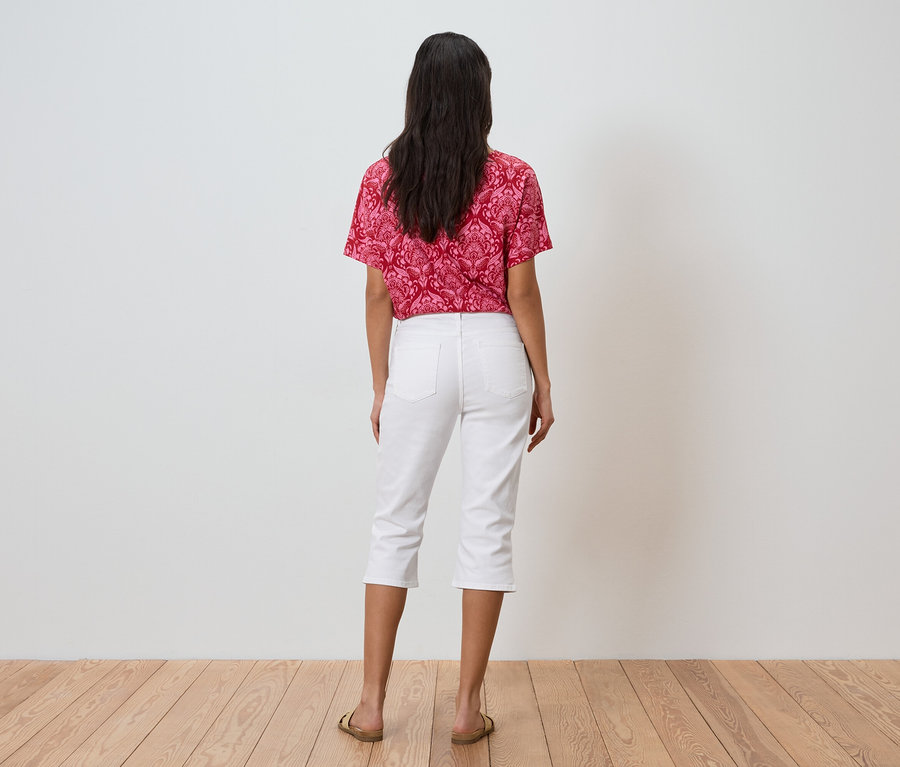 Egy sötét hajú nő áll háttal a kamerának, piros mintás felsőt, fehér háromnegyedes nadrágot és arany szandált visel.