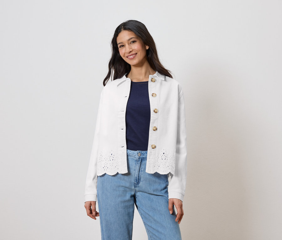 Nő visel egy fehér, lyukacsos mintázatú kabátot egy sötétkék felső fölött és világoskék farmert.
