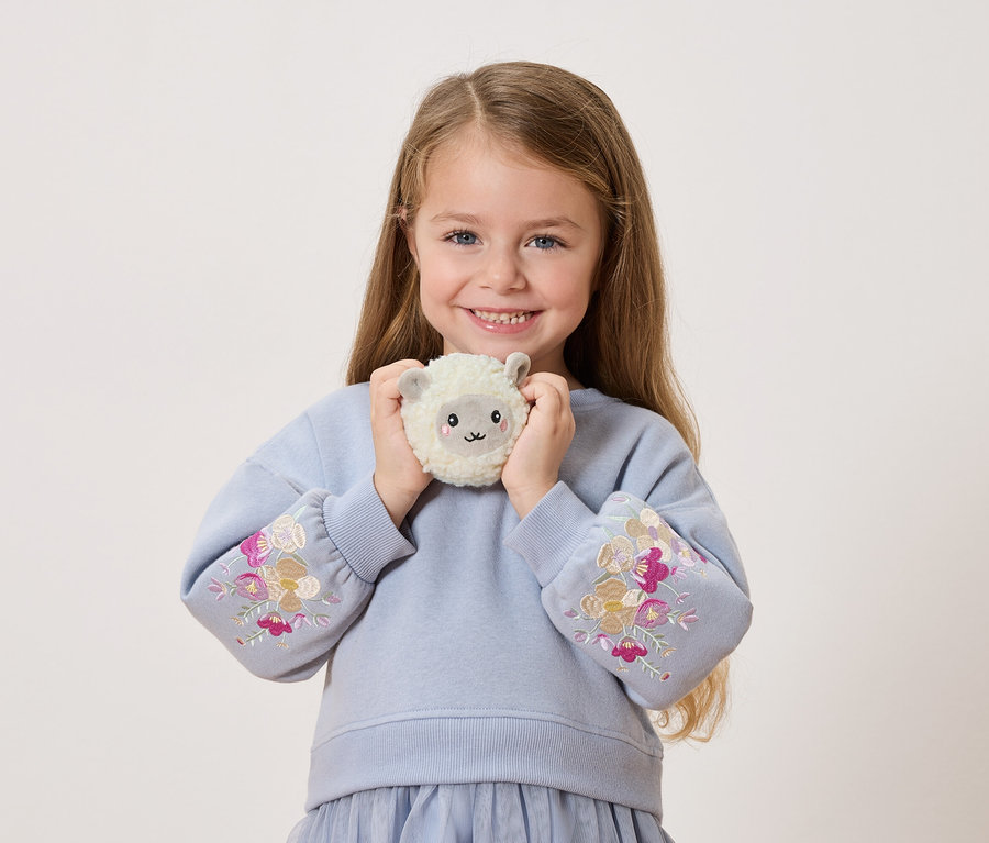 Egy mosolygó kislány egy plüss juhot tart maga előtt. Kék pulóvert visel, virágos hímzéssel az ujján.