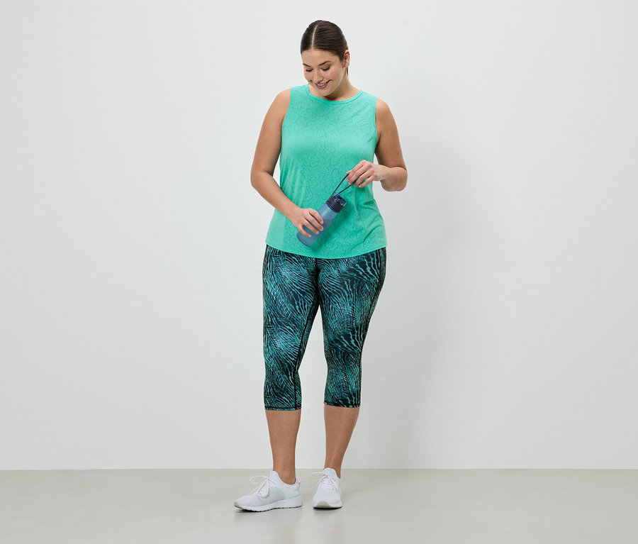 Sportruházatot viselő nő kék kulacsot tart. Zöld trikót és türkiz mintás leggingset visel.
