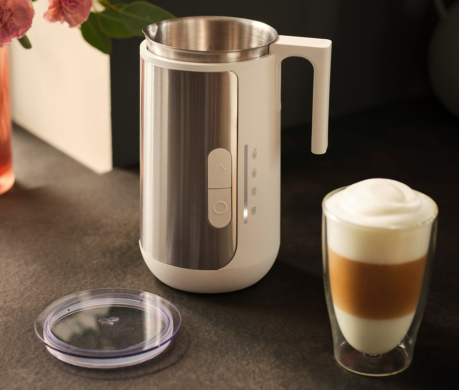 Egy rozsdamentes acélból készült tejhabosító egy latte kávéspohár és egy fedő mellett áll egy sötét pulton.