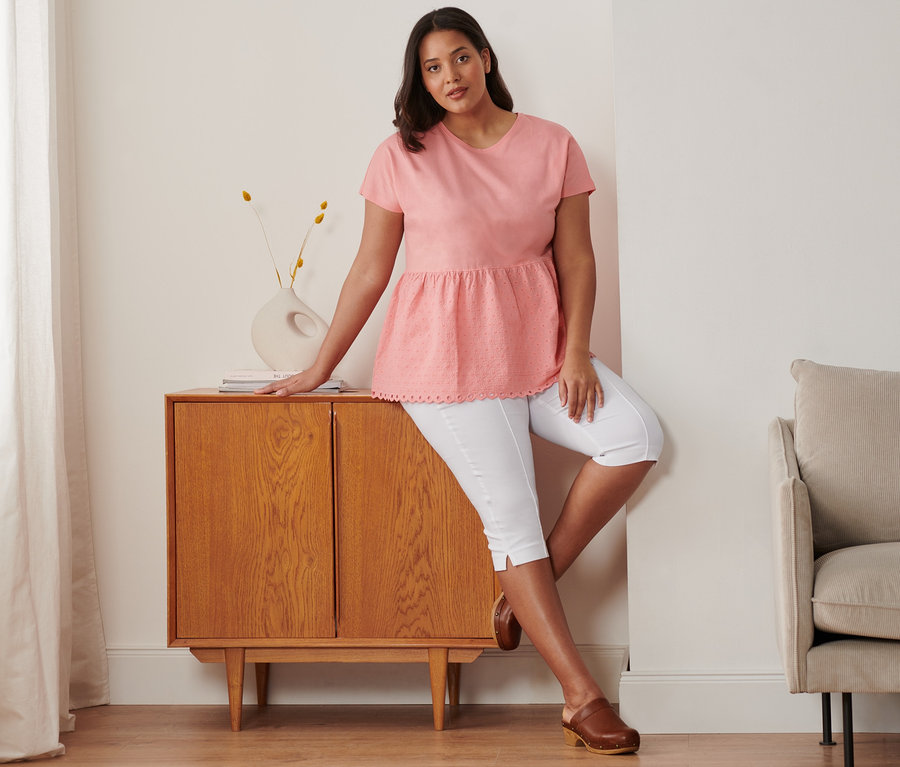 Komódon támaszkodó nő rózsaszín, lyukhímzéses felsőt, fehér, 3/4-es sztreccsnadrágot és barna bőr klumpát visel.