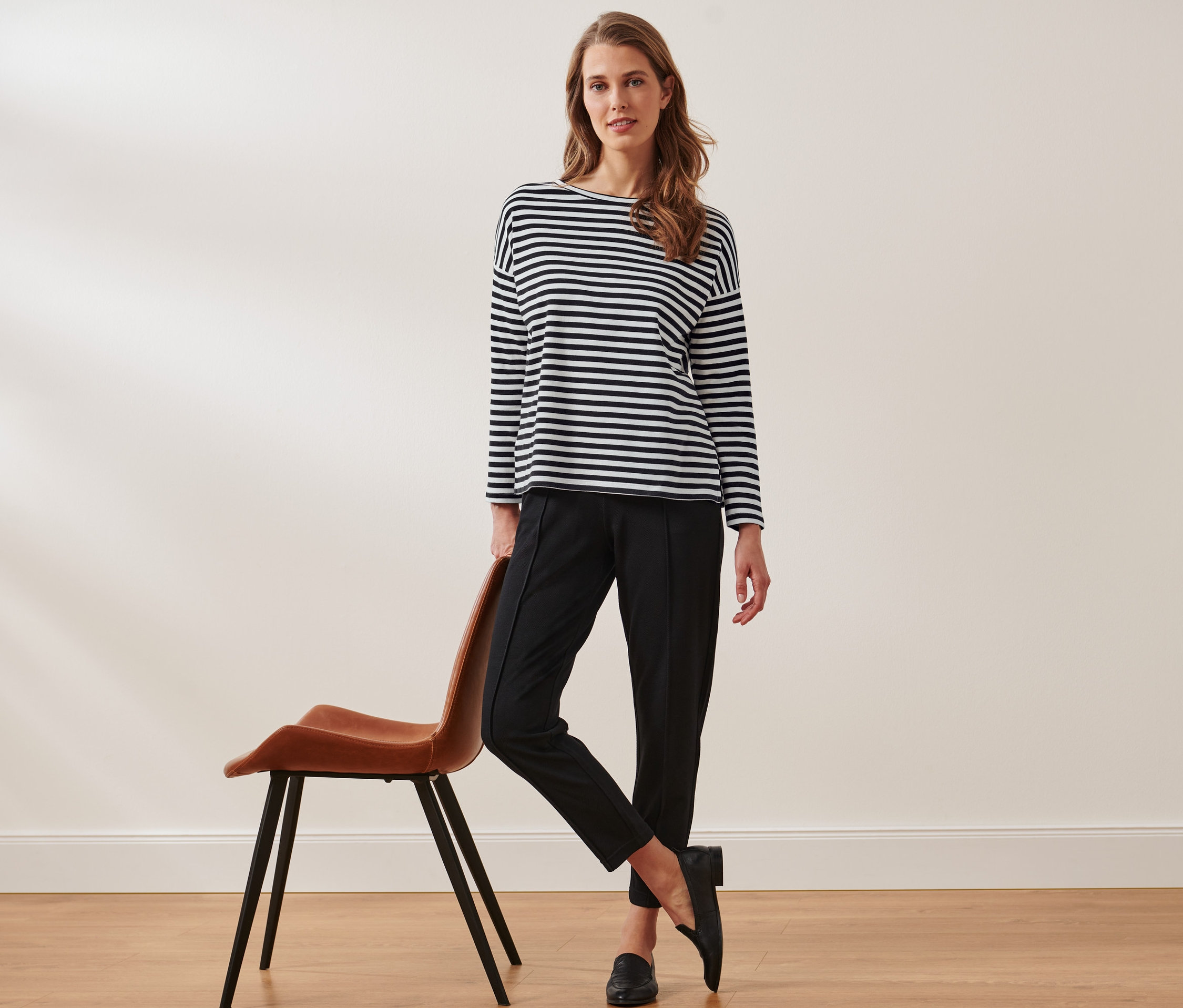 Egy nő egy barna széknek dőlve pózol fekete nadrágban és egy csíkos hosszú ujjú felsőben.