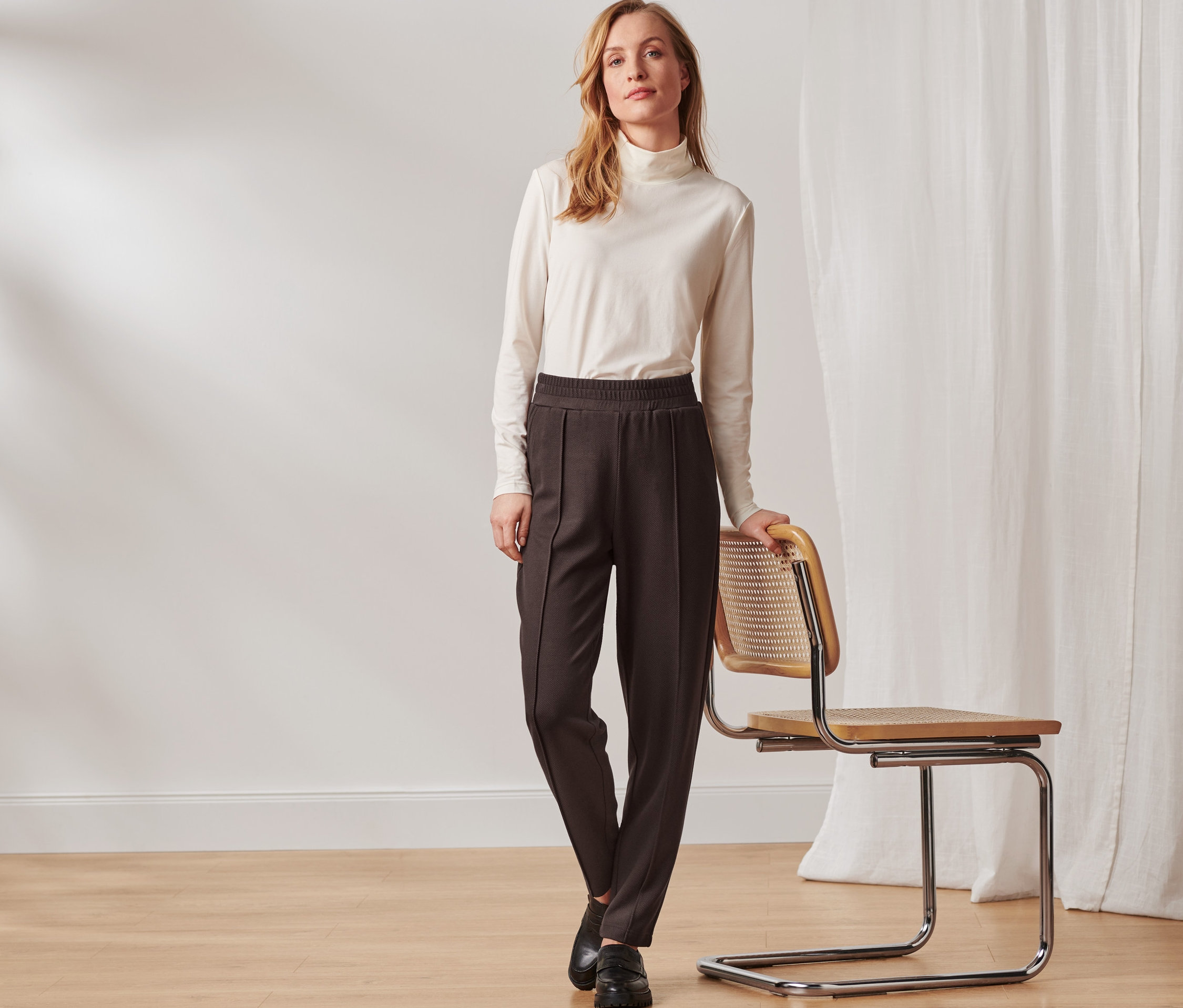 Nő áll egy szék mellett és sötétbarna, bokáig érő lezser nadrágot visel.