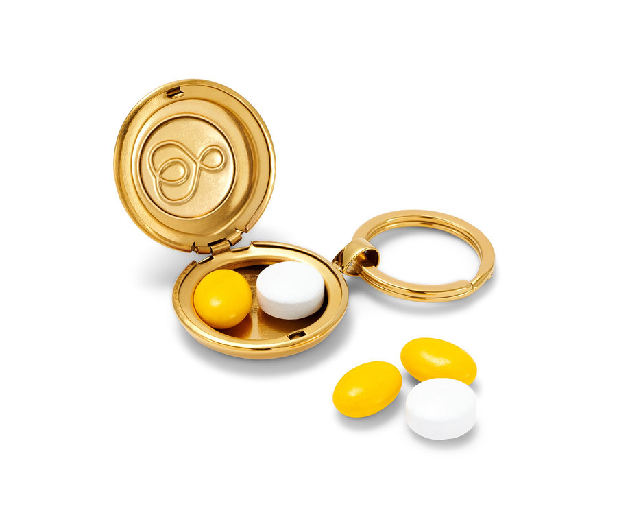 A nyitott, arany gyógyszeresdoboz-kulcstartó egy sárga és egy fehér tablettát tartalmaz, mellette két tabletta fekszik.
