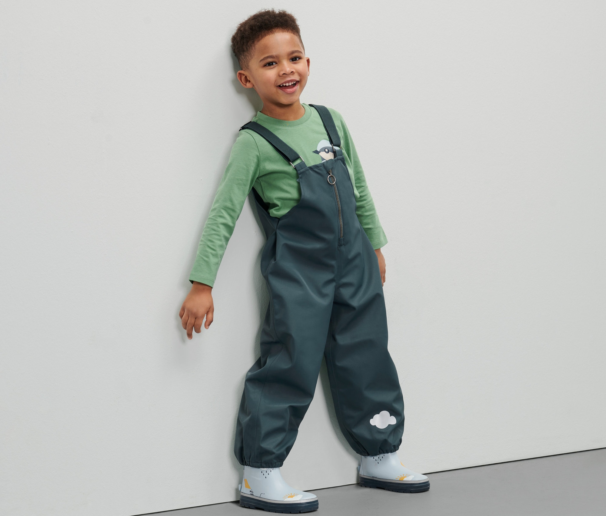 Egy kisfiú kisgyerek kantáros esőnadrágot visel és a falnak támaszkodik.