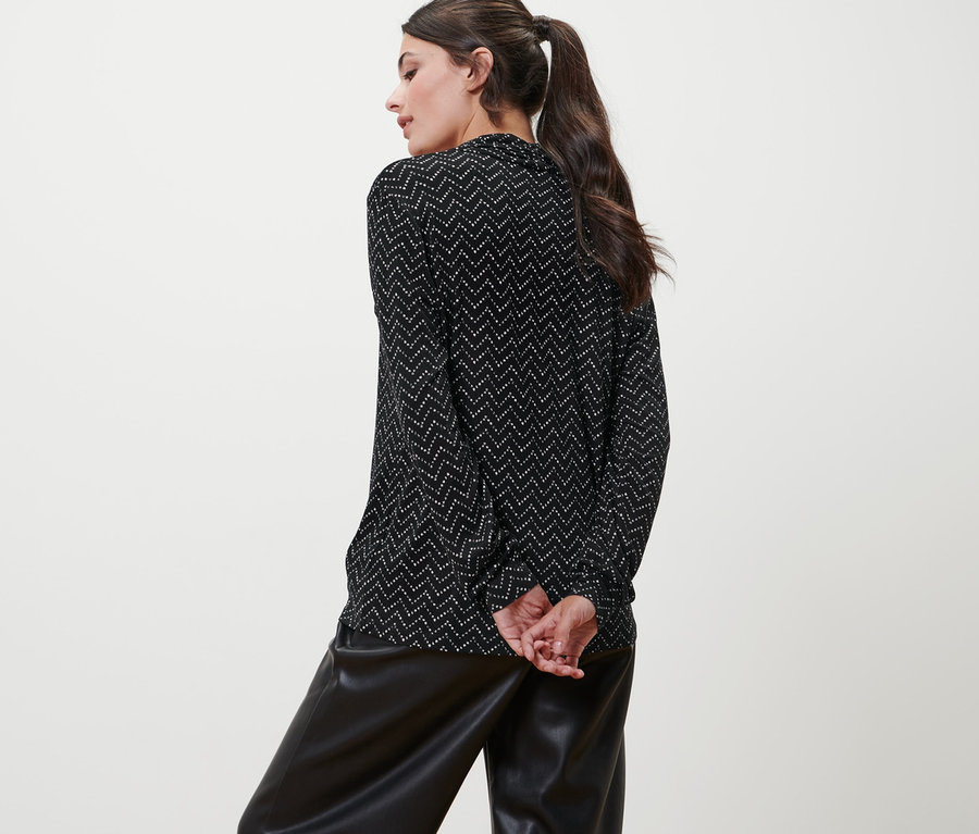 Egy nő háttal áll a kamerának, és egy nyomtatott mintás, V-nyakkivágású hosszú ujjú felsőt és egy fekete műbőr nadrágot visel.