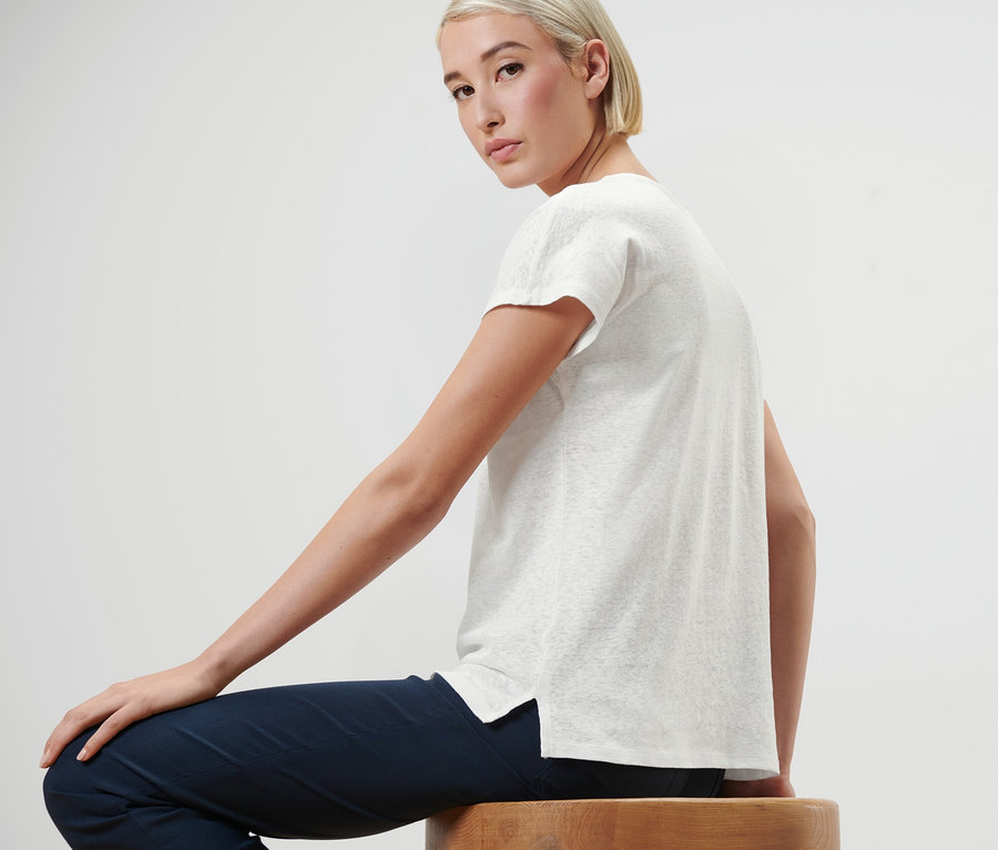 Nő ül egy fa zsámolyon, krémfehér vászon felsőt visel.