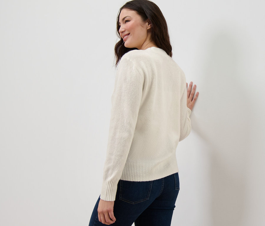 Nő néz a válla fölött, és egy krémfehér, kötött mintás kardigánban a falnak támaszkodik.