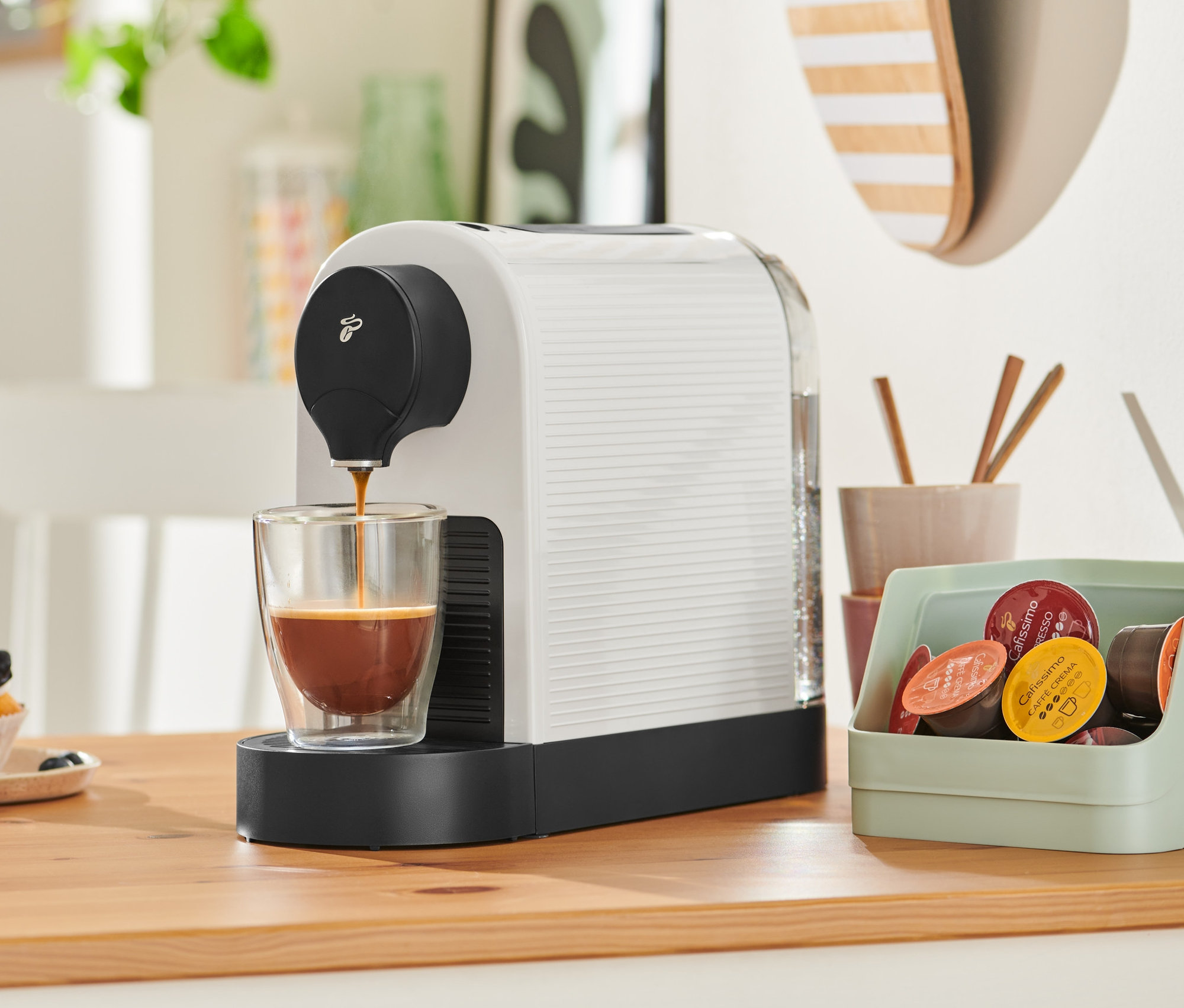 A Cafissimo pure plus, fehér kávéfőző kávét tölt egy pohárba. A háttérben egy cupcake látható.