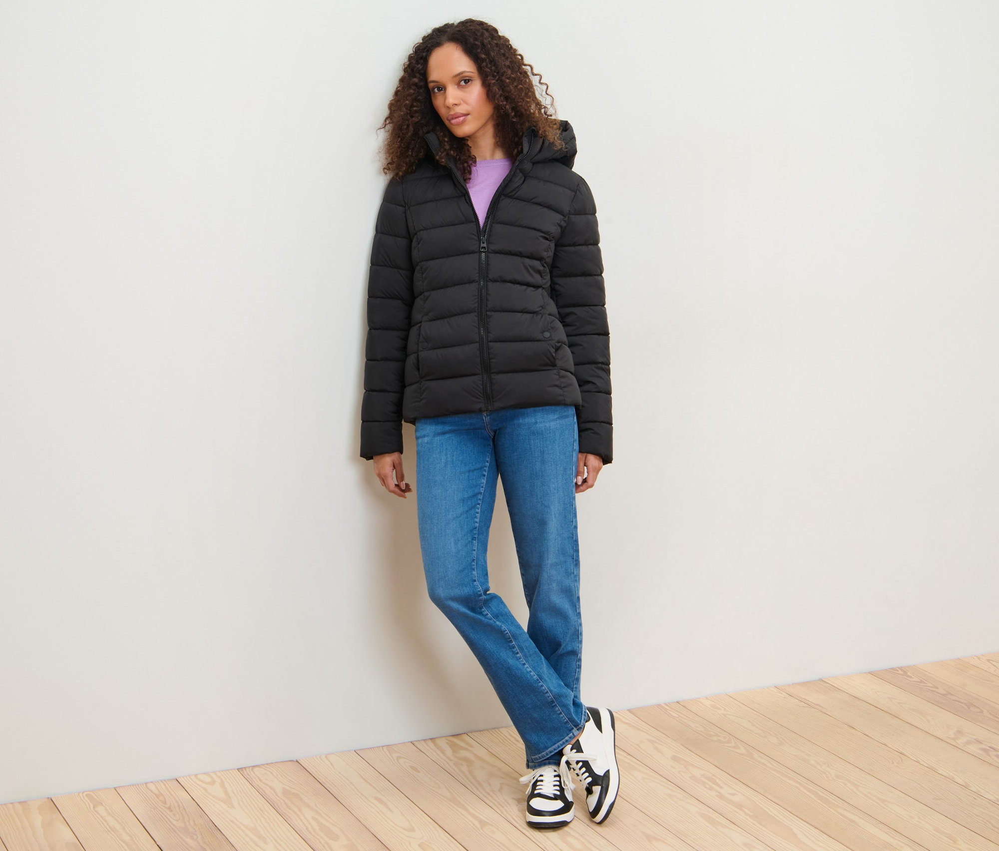 A falnak támaszkodó nő fekete steppelt dzsekit visel.