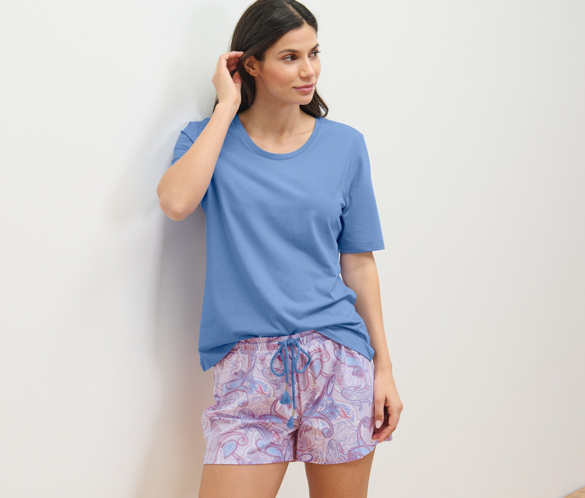 Nő rövidnadrágos pizsamában a falnak támaszkodva.